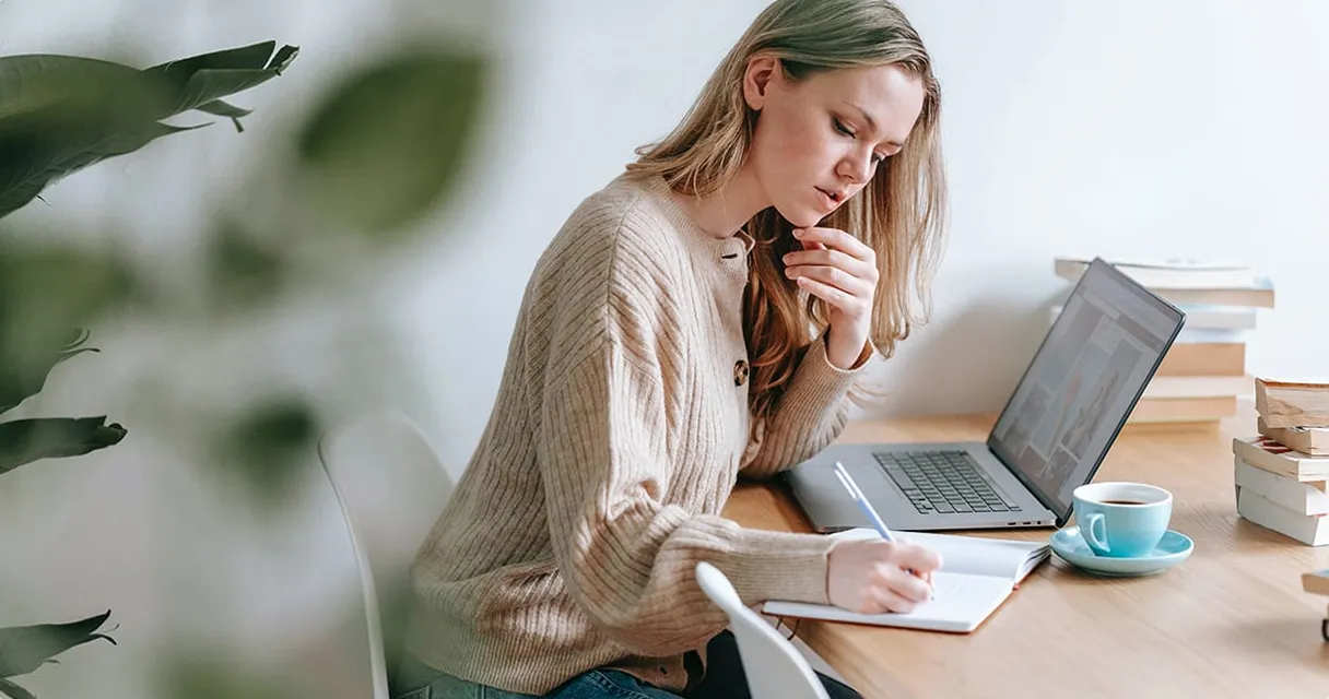 Young woman writing in a notebook at a desk with a laptop, books, and a cup of coffee. She appears focused and thoughtful.