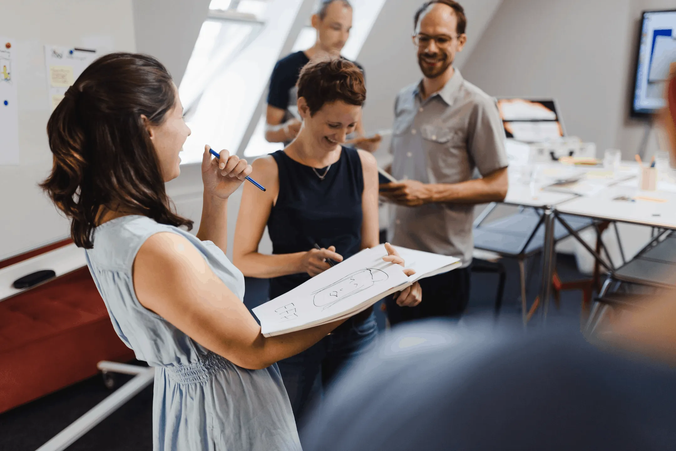 Personen stehen in einem Büro zusammen, eine hält ein Klemmbrett mit einer Skizze