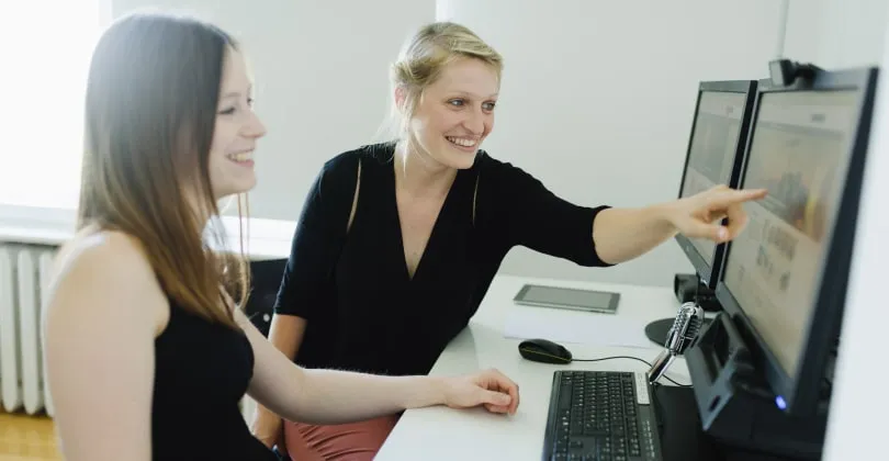 Zwei Frauen an einem Computerarbeitsplatz, eine zeigt lächelnd auf einen Monitor.
